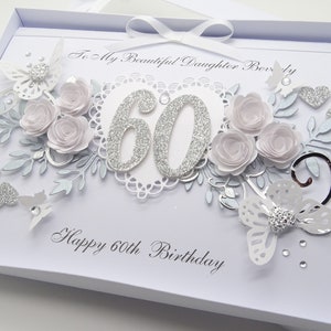 May include: A white birthday card with silver glitter accents and floral embellishments. The card features the number 60 in glitter, paper roses, butterflies, and the text "Happy 60th Birthday".