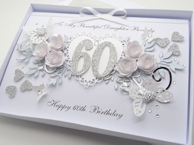 May include: A white birthday card with silver glitter accents and floral embellishments. The card features the number 60 in glitter, paper roses, butterflies, and the text "Happy 60th Birthday".
