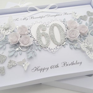 May include: A white birthday card with a decorative design. The card features the number 60 in silver glitter, surrounded by light pink paper roses, silver leaves, and glitter hearts. The text reads "Happy 60th Birthday."