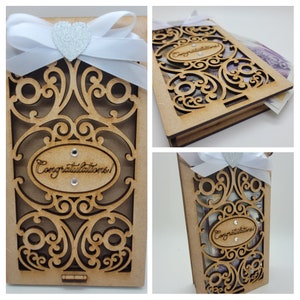 May include: A wooden gift box with an ornate, laser-cut design. The box is adorned with a white ribbon, a silver heart, and the word "Congratulations!" in an oval frame. The box is designed to hold money.