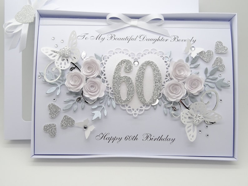 May include: A white birthday card with a decorative design. The card features the text "To My Beautiful Daughter Beverly" and "Happy 60th Birthday". Embellishments include white paper roses, butterflies, and silver glitter accents.