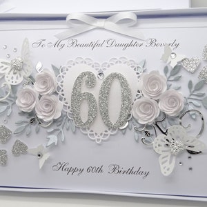 May include: A white and silver 60th birthday card with a decorative design. The card features a heart-shaped embellishment with the number 60 in silver glitter, surrounded by paper roses, butterflies, and the text "Happy 60th Birthday".