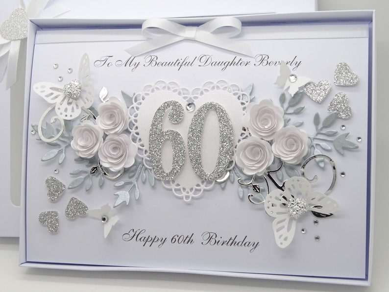 May include: A white and silver 60th birthday card with a decorative design. The card features a heart-shaped embellishment with the number 60 in silver glitter, surrounded by paper roses, butterflies, and the text "Happy 60th Birthday".