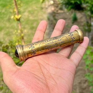 May include: A brass kaleidoscope with decorative floral patterns and the quote, "A Kaleidoscope reminds us that beauty exists in the fragments of life." The kaleidoscope has a dark lens and a gold finish.