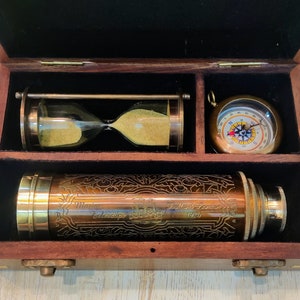 Könnte beinhalten: Holzkiste mit Messing-Teleskop, Sanduhr und Kompass. Das Teleskop ist mit den Worten "Marine Telescope" und "T. Lloyd London 1920" graviert. Die Sanduhr enthält gelben Sand. Der Kompass hat ein weißes Zifferblatt mit Richtungsmarkierungen.