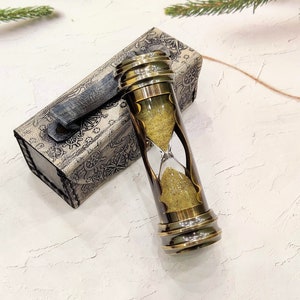 May include: An antique brass hourglass with a clear glass center filled with yellow sand. The hourglass is next to a decorative box with a nautical map design. The hourglass is approximately 6 inches tall.