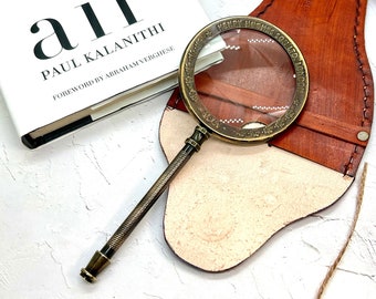 May include: A vintage-style magnifying glass with a bronze-colored frame and handle, resting on a brown leather case. The glass is positioned near a book titled "air" by Paul Kalanithi. The leather case is stitched and has a light-colored interior.