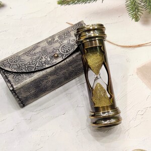 May include: A vintage brass hourglass with a sand timer, next to a matching leather case. The case is embossed with nautical symbols like anchors and compasses. The hourglass is approximately 5 inches tall.