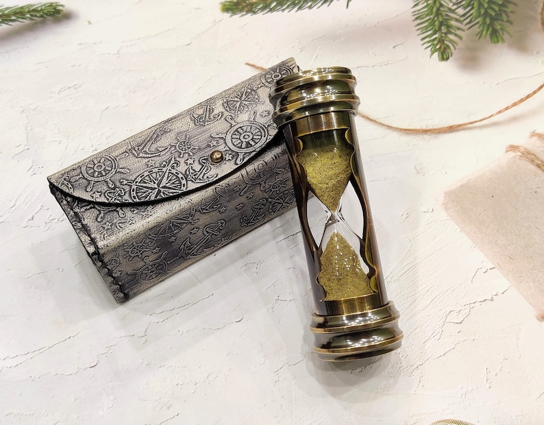 May include: A vintage brass hourglass with a sand timer, next to a matching leather case. The case is embossed with nautical symbols like anchors and compasses. The hourglass is approximately 5 inches tall.
