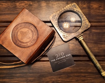 May include: A vintage-style magnifying glass with a brass handle and frame, alongside a leather-bound case. The magnifying glass has the text "HENRY HUGHES SON LTD LONDON 1941". A business card reads "THE CHOSEN ONE".