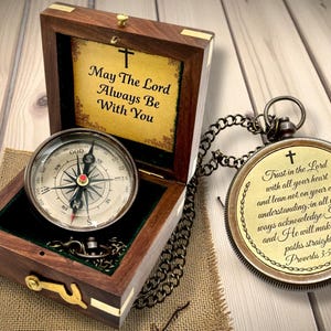 May include: A wooden box containing a compass and a pocket watch on a chain. The box displays the text "May The Lord Always Be With You" and a cross. The pocket watch has the text "Trust in the Lord" and a cross.