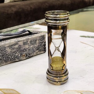 May include: An antique-style hourglass with a brass finish, filled with yellow sand. Beside it is a decorative box with nautical-themed engravings. The hourglass is a classic timekeeping device, perfect for home decor.