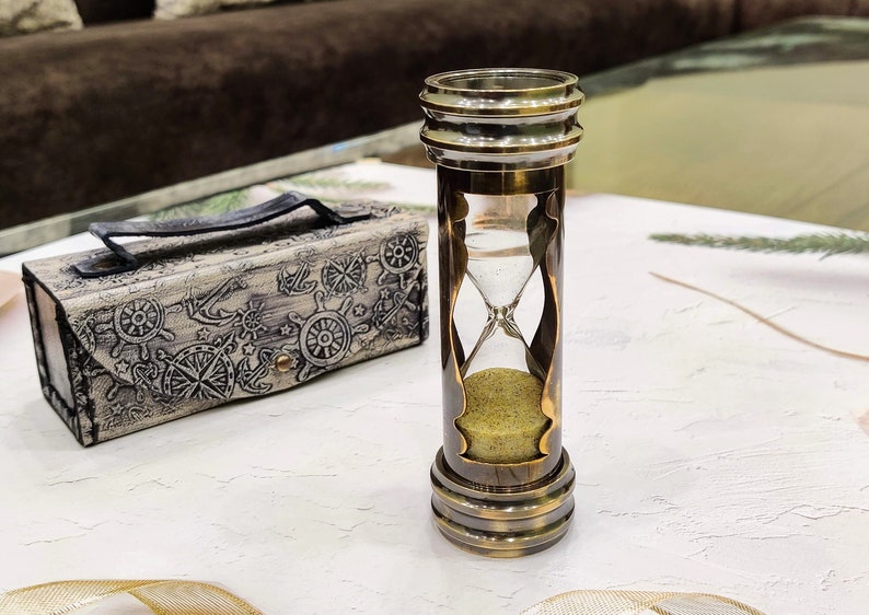 May include: An antique-style hourglass with a brass finish, filled with yellow sand. Beside it is a decorative box with nautical-themed engravings. The hourglass is a classic timekeeping device, perfect for home decor.