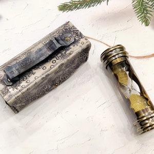 May include: An antique-style hourglass with a brass frame and a leather case. The hourglass contains yellow sand and is connected to a leather case with a handle. The case is silver with a nautical design.