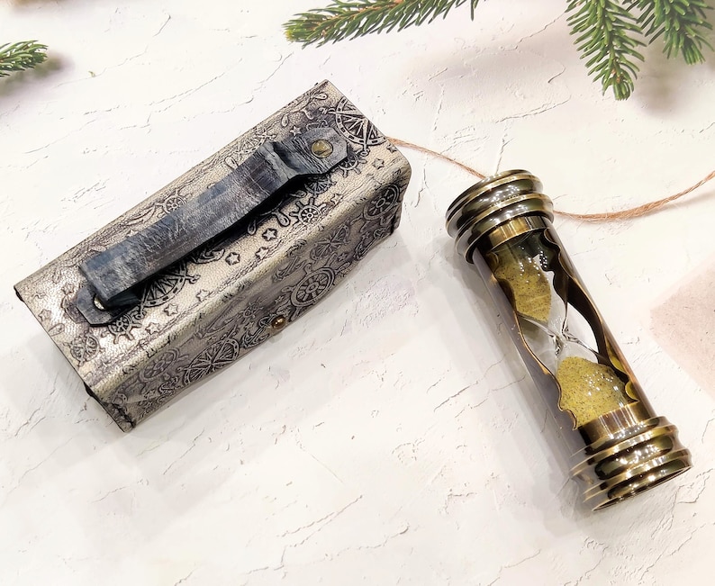 May include: An antique-style hourglass with a brass frame and a leather case. The hourglass contains yellow sand and is connected to a leather case with a handle. The case is silver with a nautical design.