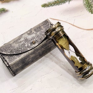 May include: An antique brass hourglass with a leather case. The hourglass features a clear glass chamber filled with yellow sand, encased in a brass frame. The case is gray with nautical-themed embossed designs.