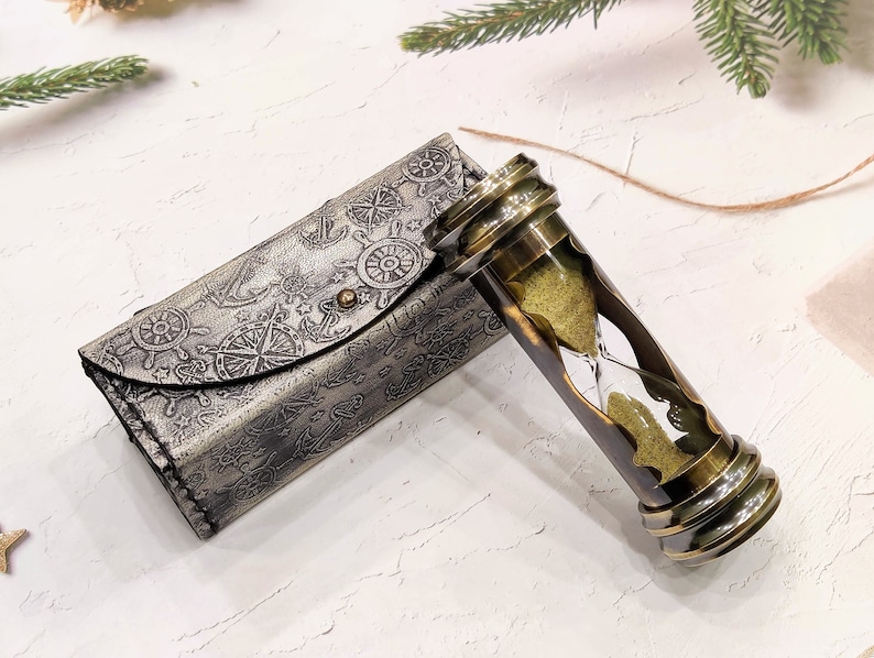 May include: An antique brass hourglass with a leather case. The hourglass features a clear glass chamber filled with yellow sand, encased in a brass frame. The case is gray with nautical-themed embossed designs.