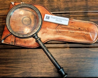 May include: A vintage magnifying glass with a detailed, ornate handle rests on a brown leather case. The magnifying glass has a silver-colored frame and a clear lens. The case is embossed with the text "HENRY HUGHES & SONS LTD. LONDON 1941". A white tag with the text "TheChosenOne Store" is also present.