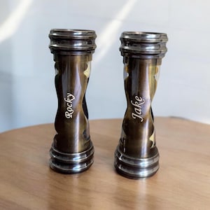 May include: Two bronze-colored, hourglass-shaped vases with a metallic finish. Each vase has a name engraved on it: "Rocky" and "Jake". The vases are displayed on a wooden surface.