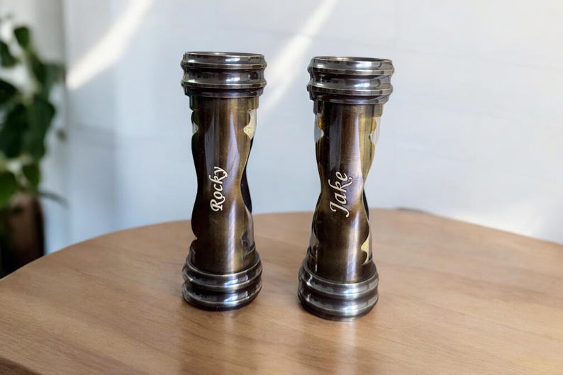 May include: Two bronze-colored, hourglass-shaped vases with a metallic finish. Each vase has a name engraved on it: "Rocky" and "Jake". The vases are displayed on a wooden surface.