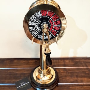 Può includere: Un telegrafo di sala macchine in ottone lucido con un quadrante in bianco e nero con indicatori di velocità e direzione. Il quadrante indica "Stop", "Avanti" e "Indietro". La base è in legno scuro e l'impugnatura è nera.