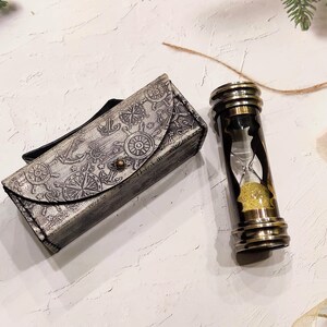 May include: An antique-style hourglass with a brass frame and a decorative leather case. The case is gray with nautical-themed embossed designs. The hourglass contains yellow sand. A star-shaped ornament is also present.