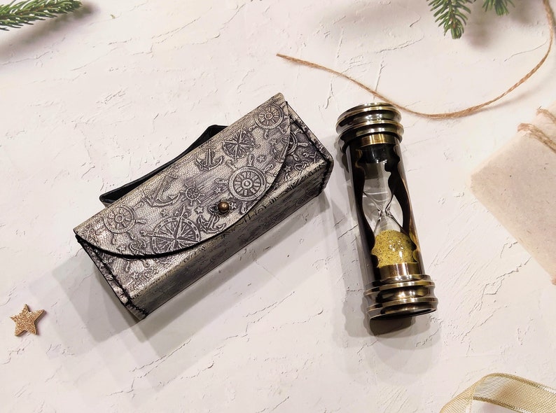 May include: An antique-style hourglass with a brass frame and a decorative leather case. The case is gray with nautical-themed embossed designs. The hourglass contains yellow sand. A star-shaped ornament is also present.