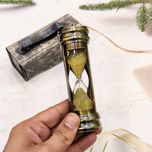 May include: A brass hourglass with a sand timer, held by a hand. The hourglass is cylindrical with a clear glass center and brass ends. A decorative box with a handle is in the background. The sand is a light yellow color.