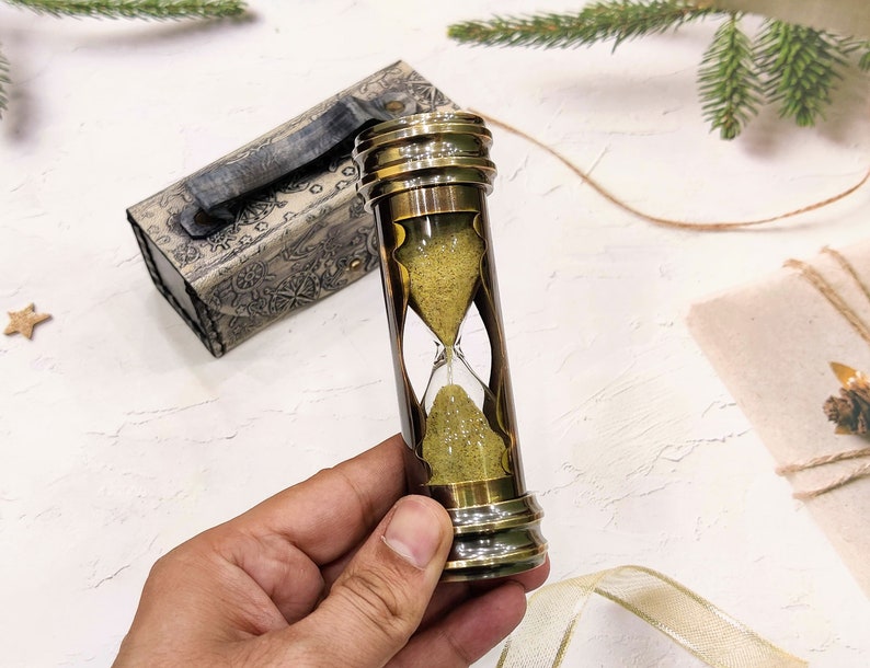 May include: A brass hourglass with a sand timer, held by a hand. The hourglass is cylindrical with a clear glass center and brass ends. A decorative box with a handle is in the background. The sand is a light yellow color.