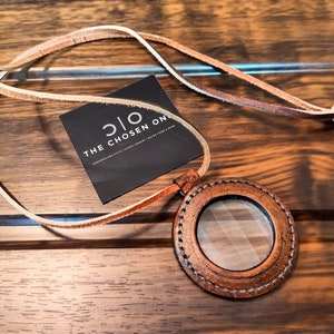 Puede incluir: Un collar con colgante de cuero marrón con un centro de cristal transparente. El colgante es circular con detalles cosidos y un cordón de cuero. También hay una etiqueta negra con el texto blanco "THE CHOSEN ONE".