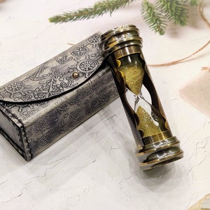 May include: An hourglass with a brass frame and a dark brown center, filled with yellow sand. It sits next to a matching case with nautical-themed embossed designs. The case has a button closure.