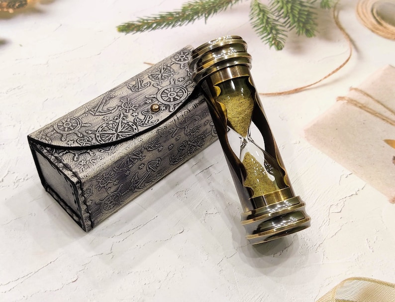 May include: An hourglass with a brass frame and a dark brown center, filled with yellow sand. It sits next to a matching case with nautical-themed embossed designs. The case has a button closure.