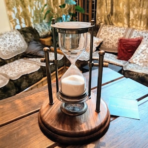 May include: An hourglass with a wooden base and metal frame. The glass chamber contains white sand. A compass design is visible on the base. The hourglass is on a wooden table. A card with the text "THE CHOSEN ONE" is on the table.