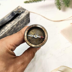 May include: A vintage-style brass compass with a detailed face, held in a hand. The compass features directional markings and a needle. A decorative box with a patterned design is in the background.