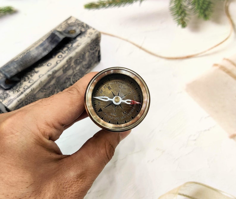 May include: A vintage-style brass compass with a detailed face, held in a hand. The compass features directional markings and a needle. A decorative box with a patterned design is in the background.