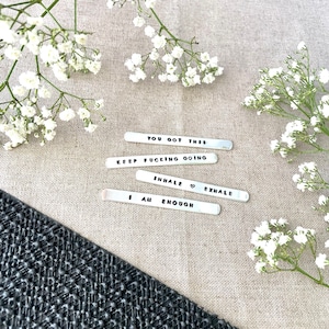 May include: Four silver metal tags with motivational phrases: "You got this", "Keep fucking going", "Inhale ♥ Exhale", and "I am enough". The tags are on a light brown fabric background with white flowers.