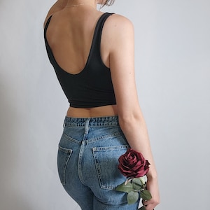 K&ouml;nnte beinhalten: Eine Frau tr&auml;gt ein schwarzes Tanktop und eine blaue Jeans und h&auml;lt eine einzelne rote Rose.