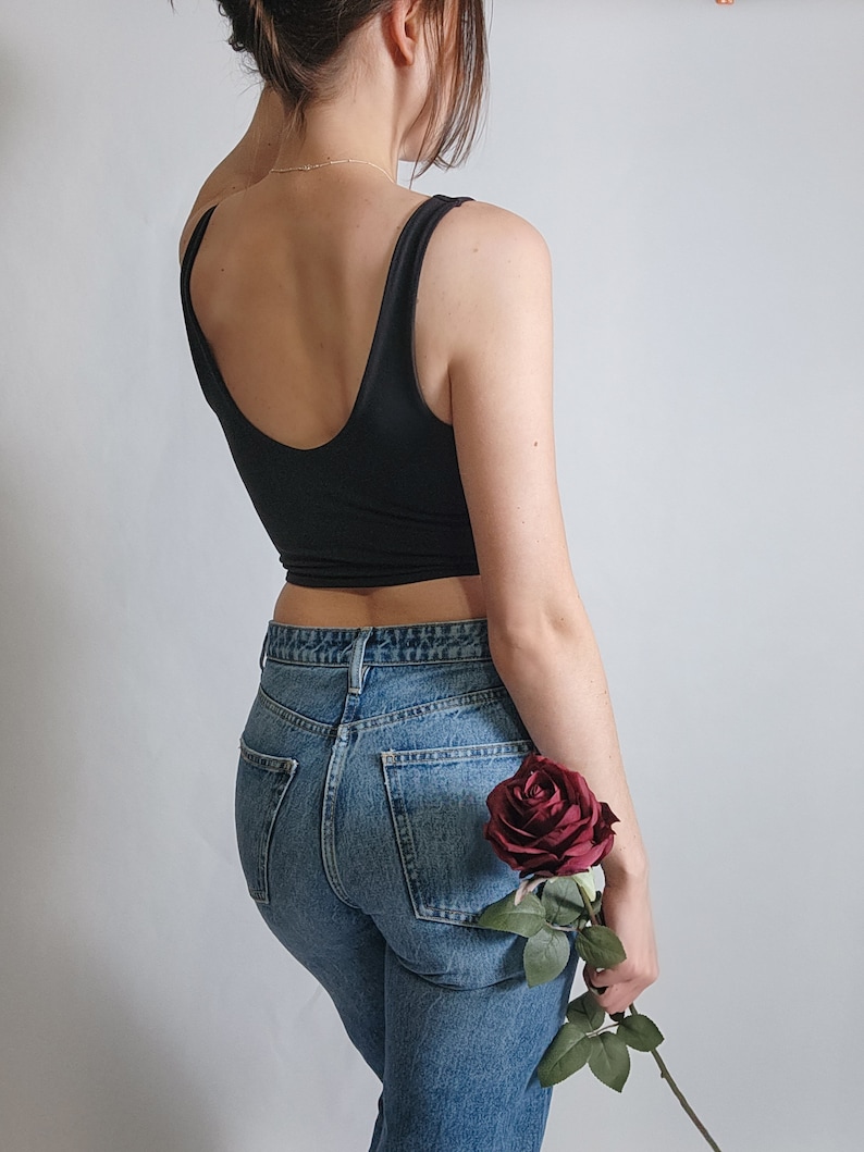 K&ouml;nnte beinhalten: Eine Frau tr&auml;gt ein schwarzes Tanktop und eine blaue Jeans und h&auml;lt eine einzelne rote Rose.