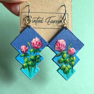 May include: A pair of painted wooden earrings featuring a blue and green colour scheme with a design of pink clover flowers. The earrings are attached to gold hooks.