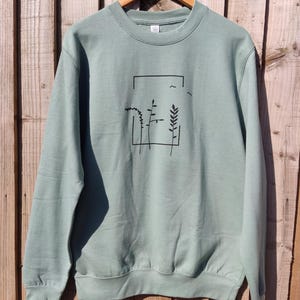 May include: Sage green sweatshirt with a crew neck and long sleeves. The front features a black graphic of plants and birds within a rectangular frame. The sweatshirt is hanging on a wooden hanger against a wooden fence background.