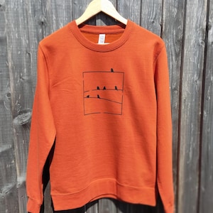 Op de afbeelding: Een roestoranje sweater met ronde hals en een zwarte grafische print op de borst. Het ontwerp bestaat uit een vierkant kader met drie horizontale lijnen en kleine zwarte vogel-silhouetten die op de draden zitten.