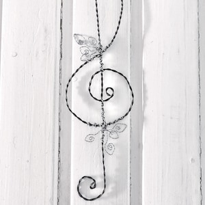 Könnte beinhalten: Ein schwarzer Notenschlüssel aus Draht mit floralen Akzenten ist auf einer weißen Holzoberfläche montiert. Das Musiksymbol ist aus verdrilltem Draht gefertigt, mit zarten Drahtblumen, die Details hinzufügen. Der Hintergrund ist eine weiße Wand mit vertikalen Brettern.