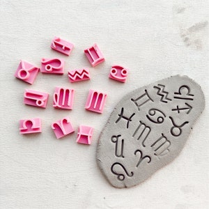Op de afbeelding: Roze rubberen stempels met astrologische symbolen, gerangschikt op een wit oppervlak. Een grijze klei-plaat met de astrologische symbolen erop gedrukt.
