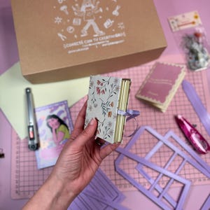 以下が含まれることがあります： 「Box Creativa」の文字とイラストが入ったクラフトボックス。紫のリボンが付いた小さな花柄のノートが手に持たれています。その他のクラフト用品には、ピンクのカッティングマット、ホッチキス、紫色のステンシルが含まれています。