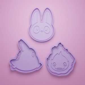 Pode incluir: Três cortadores de biscoitos roxos claros em forma de coelho, cabeça de coelho e personagem de desenho animado com chamas. Os cortadores de biscoitos estão em um fundo rosa claro.