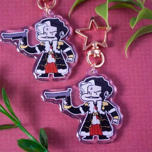 May include: Two clear acrylic keychains featuring a cartoon character with black and white hair, wearing a black and gold coat, red trousers, and holding a gun. The character has a mischievous expression. The keychains are attached to gold star-shaped keyrings.