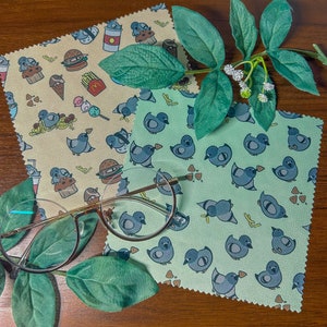 Op de afbeelding: Twee groene microvezel schoonmaakdoekjes met een herhalend patroon van cartoon duiven. De doekjes zijn gevouwen en liggen op een houten oppervlak met een bril erop.