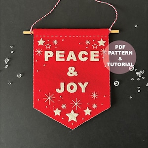 May include: A red felt banner with silver lettering spelling "PEACE & JOY", decorated with embroidered stars and snowflakes. It hangs from a wooden dowel and a red and white string. The text "PDF PATTERN & TUTORIAL" is also visible.