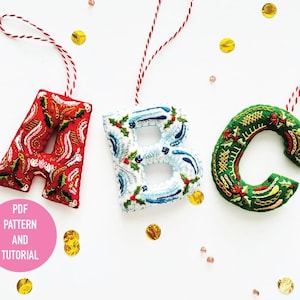 May include: Three felt ornaments shaped like the letters A, B, and C. Each letter is embroidered with colorful patterns and has a red string attached for hanging. The letters are arranged in a row on a white background with gold and pink confetti scattered around them. The text "PDF PATTERN AND TUTORIAL" is written in pink on a white circle.