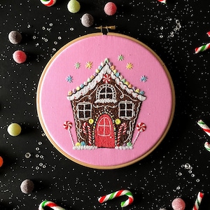 May include: Embroidered gingerbread house on a pink background in a wooden embroidery hoop. The hoop is surrounded by candy canes and gumdrops on a black background.